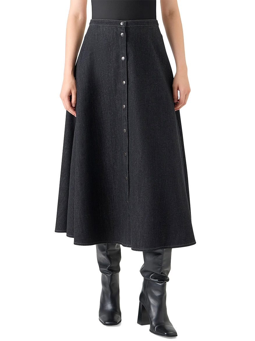 A person wears the Akris Punto Stretch Denim Snap Button Midi Skirt in Black Denim with black knee-high boots and a cotton poplin shirt, standing against a white background.