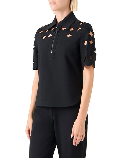 A woman wears the Akris Punto Dropping Dot Cut Out Polo in black, featuring geometric details on the shoulders and sleeves, paired with black pants.