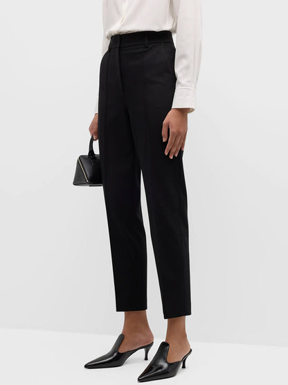 A person stands against a plain white background, wearing a white shirt, black Akris Punto Elements Ferry Tapered Jersey Pants in Black, black heeled shoes, and holding a small black handbag.