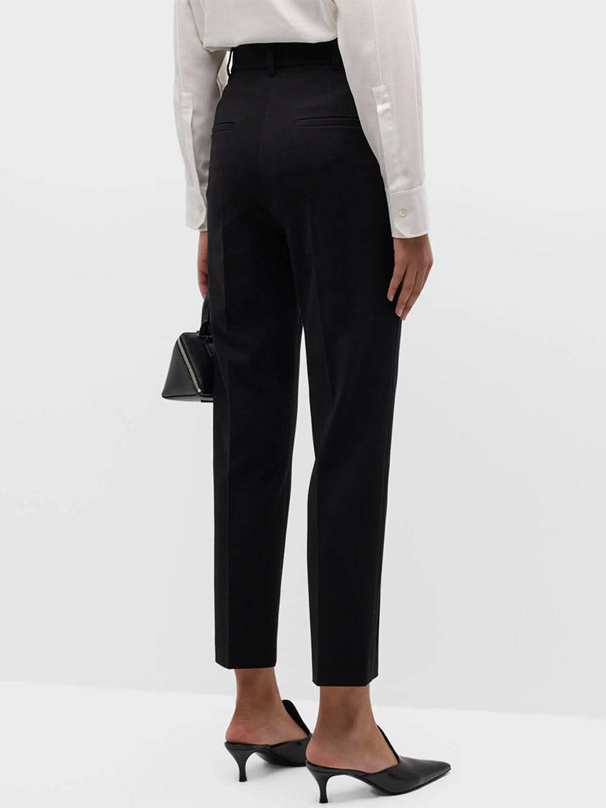 Rear view of a person wearing Akris Punto Elements Ferry Tapered Jersey Pants in Black, a white long-sleeve shirt, black heeled mules, and holding a small black handbag against a plain background.