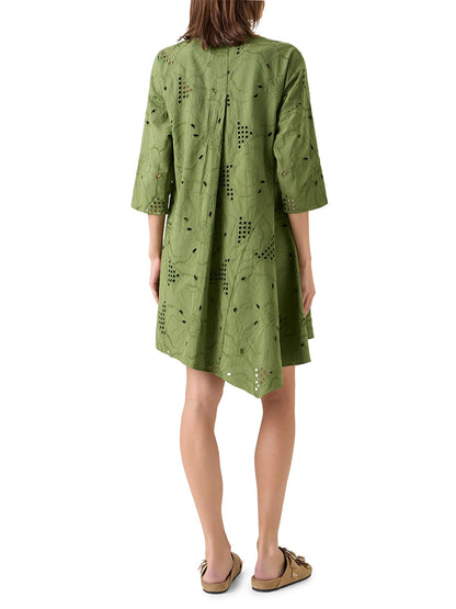 A woman stands facing away, wearing the Akris Punto Flower Eyelet Embroidered Handkerchief Dress in Green Tea and tan slip-on sandals.