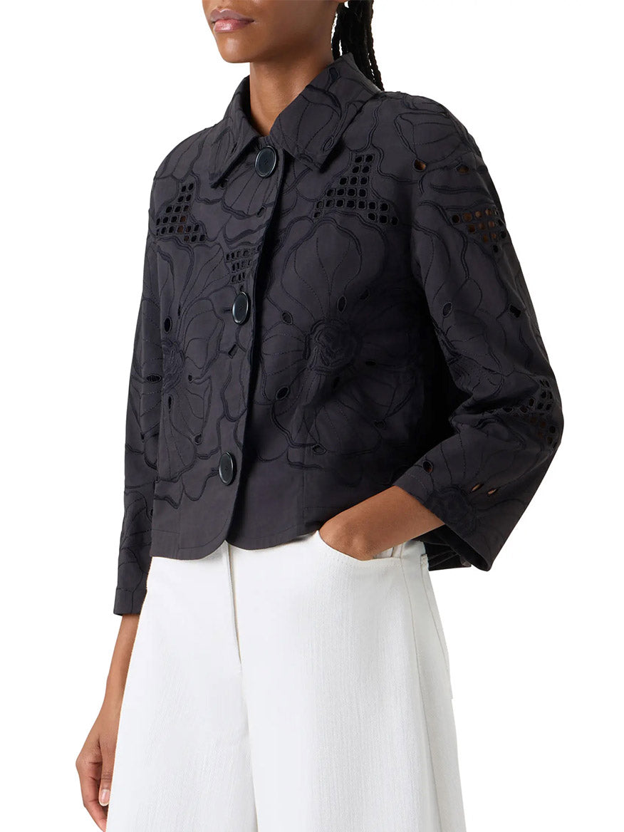 Person shown from shoulders to hips wearing the Akris Punto Flower Eyelet Embroidered Jacket in black, featuring oversized floral embroidery and large buttons, paired with white pants.