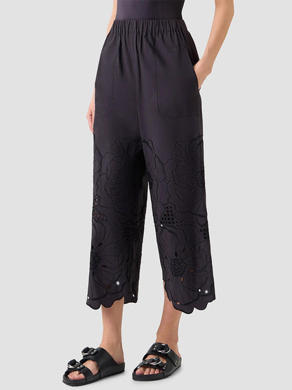 Someone stands against a plain background wearing Akris Punto Frey Flower Eyelet Embroidered Pants in black, featuring cropped cotton poplin with floral embroidery, paired with black slide sandals.