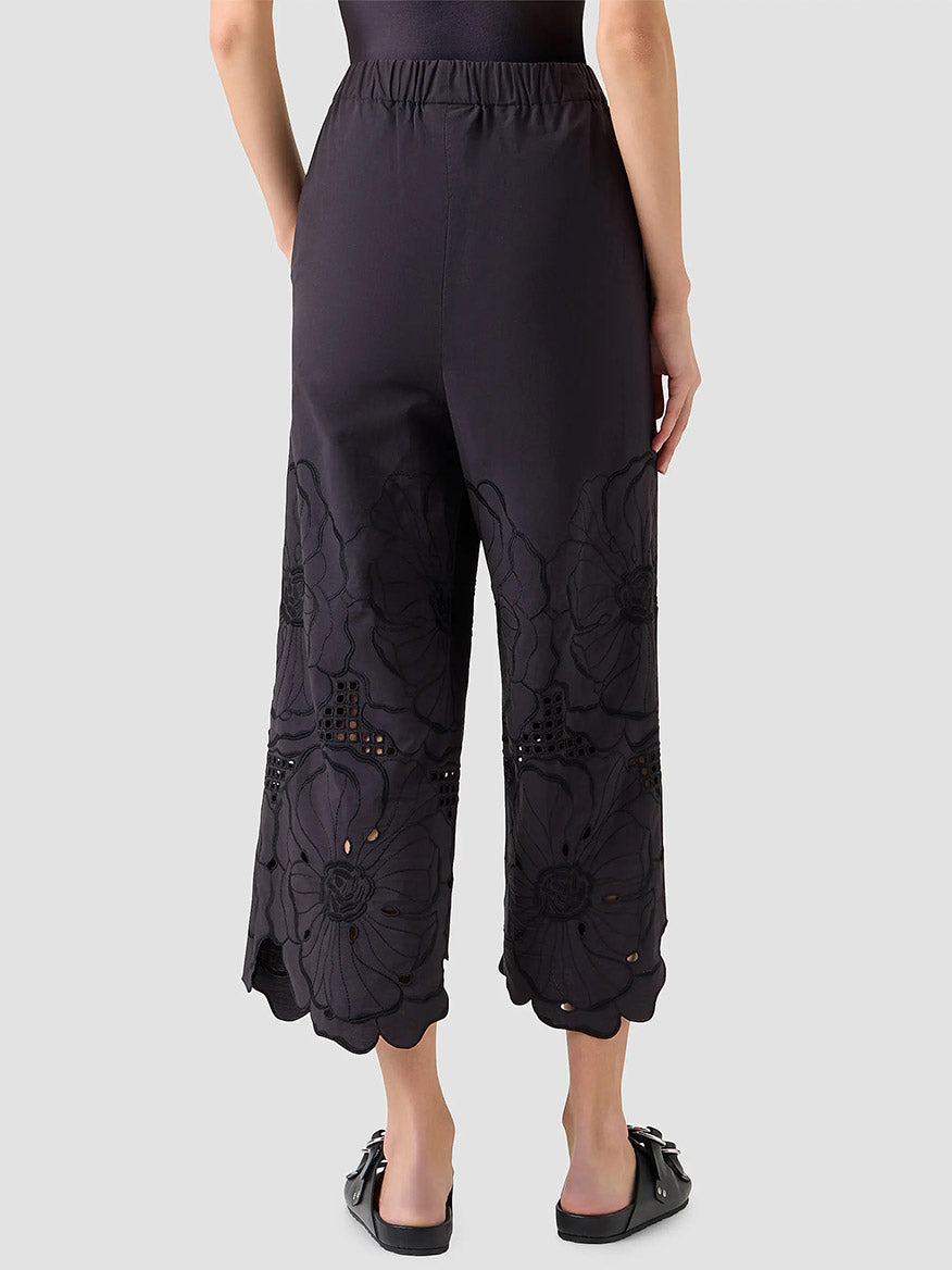 A person stands with their back to the camera, showcasing the cropped length of the Akris Punto Frey Flower Eyelet Embroidered Pant in Black, which features floral embroidery near the hems against a plain background.