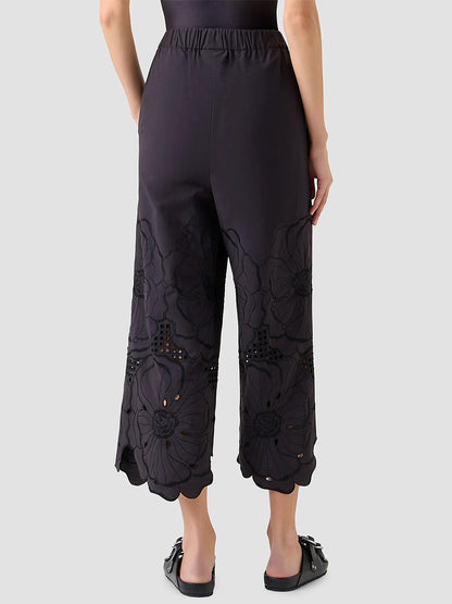 A person stands with their back to the camera, showcasing the cropped length of the Akris Punto Frey Flower Eyelet Embroidered Pant in Black, which features floral embroidery near the hems against a plain background.