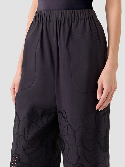A person wears the Akris Punto Frey Flower Eyelet Embroidered Pant in Black—loose-fitting cotton poplin pants with front pockets and floral embroidery near the knees, shown from waist to mid-thigh.