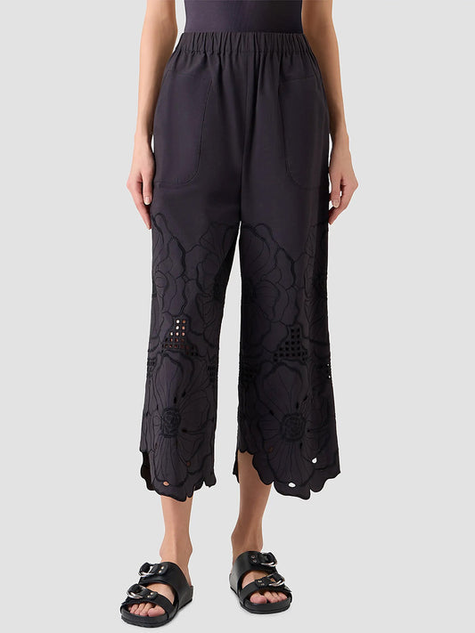 A person stands against a plain background wearing the Akris Punto Frey Flower Eyelet Embroidered Pant in Black, featuring floral cut-out designs near the hem. The cropped length and black sandals highlight its modern, intricate details.