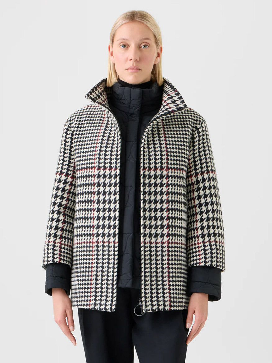 A person with straight blond hair and a neutral expression wears the Akris Punto Glencheck 2-in-1 Car Coat in Black/Cream/Vermillion over a black quilted jacket, standing forward.