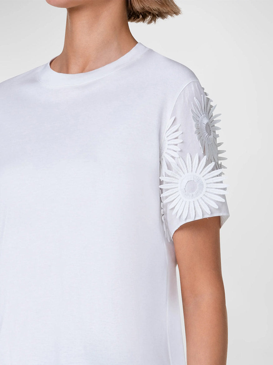 A close-up of a person wearing a white cotton jersey t-shirt with embroidered floral details on the sleeve by Akris Punto Hello Sunshine Embroidered Sleeve Tee in Cream.