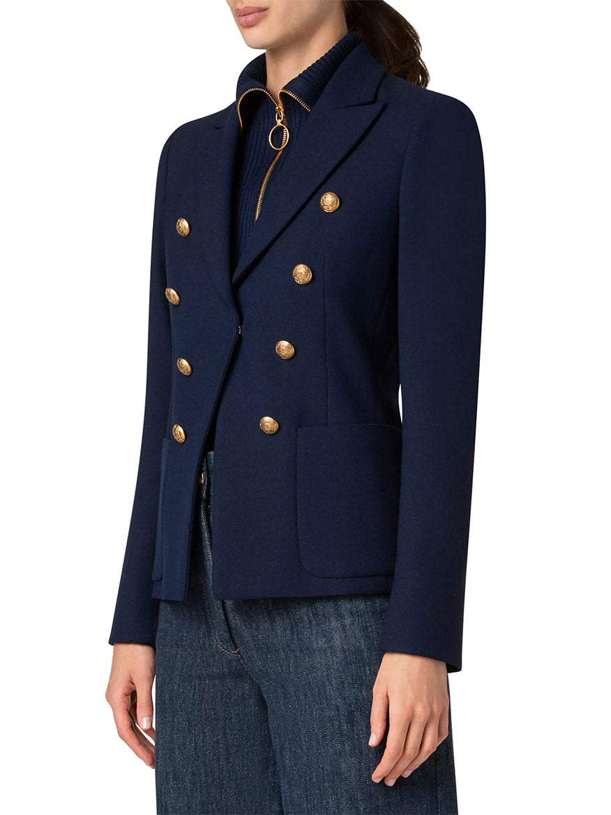 A person wears the Akris Punto Illusion Peak Lapel Blazer with gold buttons in navy over a dark top with a visible zipper, paired effortlessly with jeans.