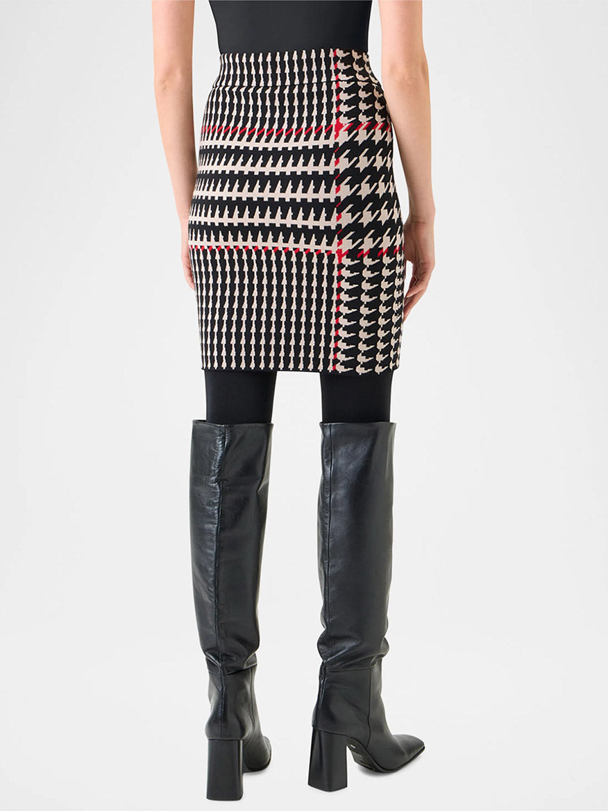 A person is seen from the back wearing a black top, black tights, knee-high black boots, and an Akris Punto Fitted Wool-Blend Plaid Jacquard Skirt in Black/Cream/Vermillion.