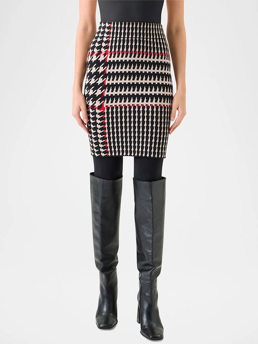 A person wearing a black top, black tights, knee-high black boots, and the Akris Punto Fitted Wool-Blend Plaid Jacquard Skirt in Black/Cream/Vermillion featuring a houndstooth pattern with red accents and a high-rise waistband.