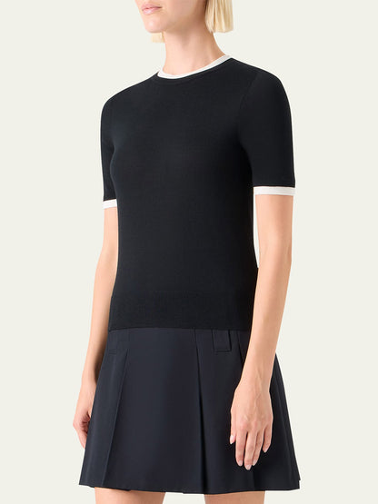 A woman stands against a plain background wearing the Akris Punto Layered Short-Sleeve Knit Sweater in Black/Cream, featuring white trim at the collar and sleeves, paired with a black pleated skirt.