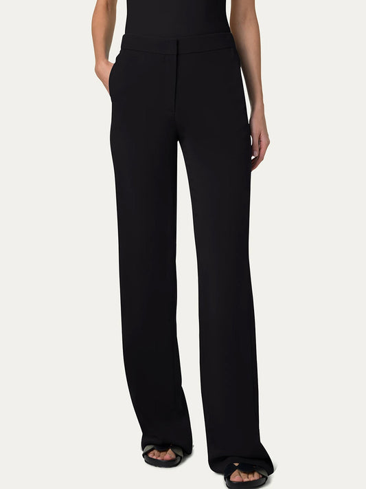 A person wearing Akris Punto Marla Wide Straight Lasercut Trousers in black, featuring an elastic waist and side slash pockets, along with black open-toe sandals and hands casually in pockets against a plain background.