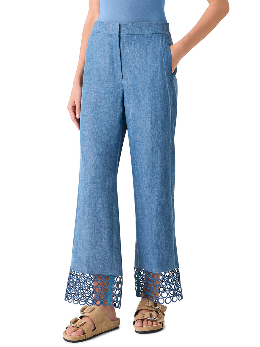 Person wearing Akris Punto Marlon Washed Denim Circle Lace Pants in Medium Blue featuring signature dot motif and circular cutout hem, paired with a light blue top and tan sandals. Hands are in pockets.