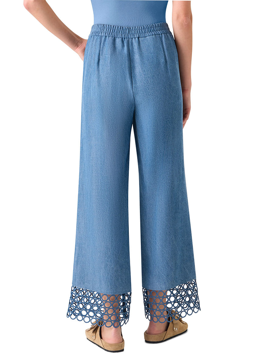 A person is shown from the back wearing Akris Punto Marlon Washed Denim Circle Lace Pants in medium blue, featuring wide legs, an elastic waistband, and open-circle lace cut-outs at the hem, paired with tan moccasin-style shoes.