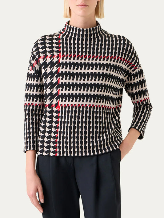 A woman wears the Akris Punto Mock Neck Plaid Sweater in Black/Cream/Vermillion, featuring a mock neckline and long sleeves with a geometric pattern, paired with dark trousers.