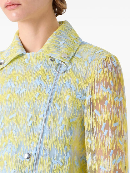 A person shown from the shoulders up wears the Akris Punto Scribble Dot Embroidery Biker Jacket in Lemon Zing/Sky Blue, featuring semi-sheer fabric, scribble dot embroidery, zippers, and metal snap buttons.
