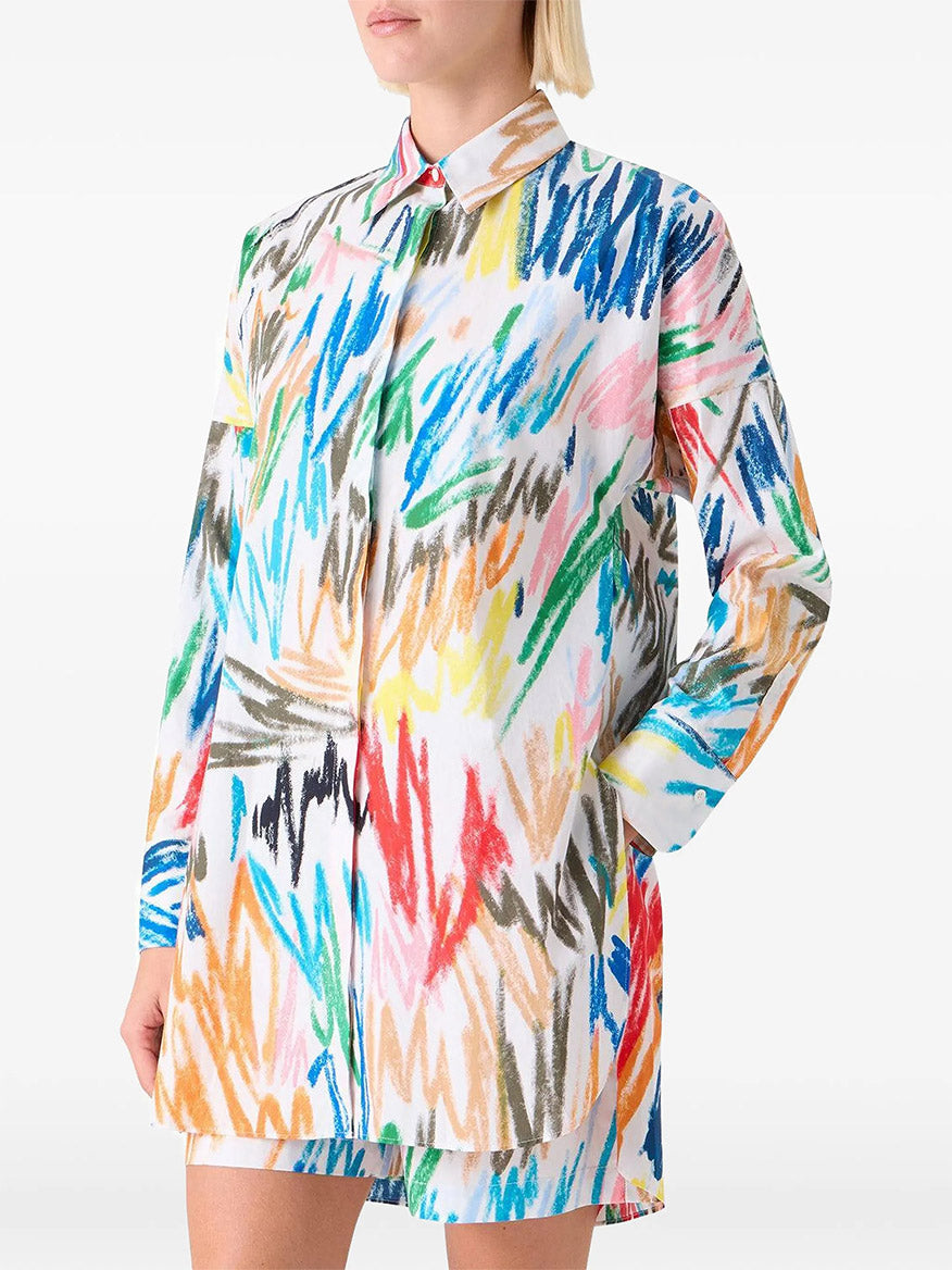 A person wearing the Akris Punto Scribble Print Tunic Blouse in Cream Multi with long sleeves and shorts, featuring a colorful abstract scribble pattern, stands with one hand in their pocket.