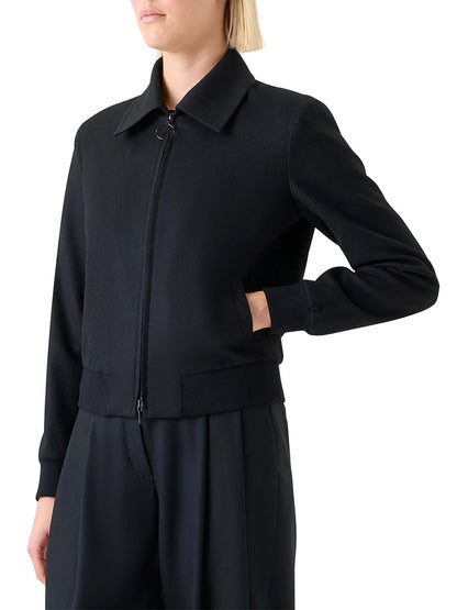 A person wears the Akris Punto Wool/Cashmere Short Bomber Jacket in Black, featuring rib-knit trim, standing with one hand in a pocket against a plain background.