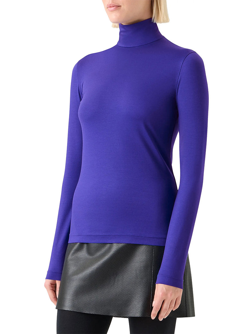 A woman wearing the Akris Punto Mock Neck Signature Dot Jersey Turtleneck in Ultra Violet, paired with a black textured mini skirt and black leggings, stands against a white background.