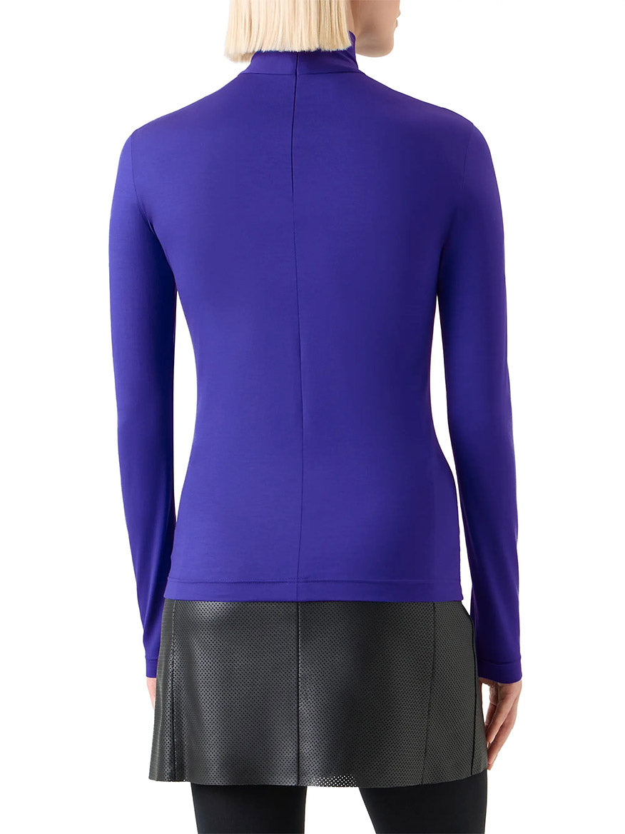 A person with light blonde hair, seen from behind, wears the Akris Punto Mock Neck Signature Dot Jersey Turtleneck in Ultra Violet paired with a black textured skirt and black leggings.