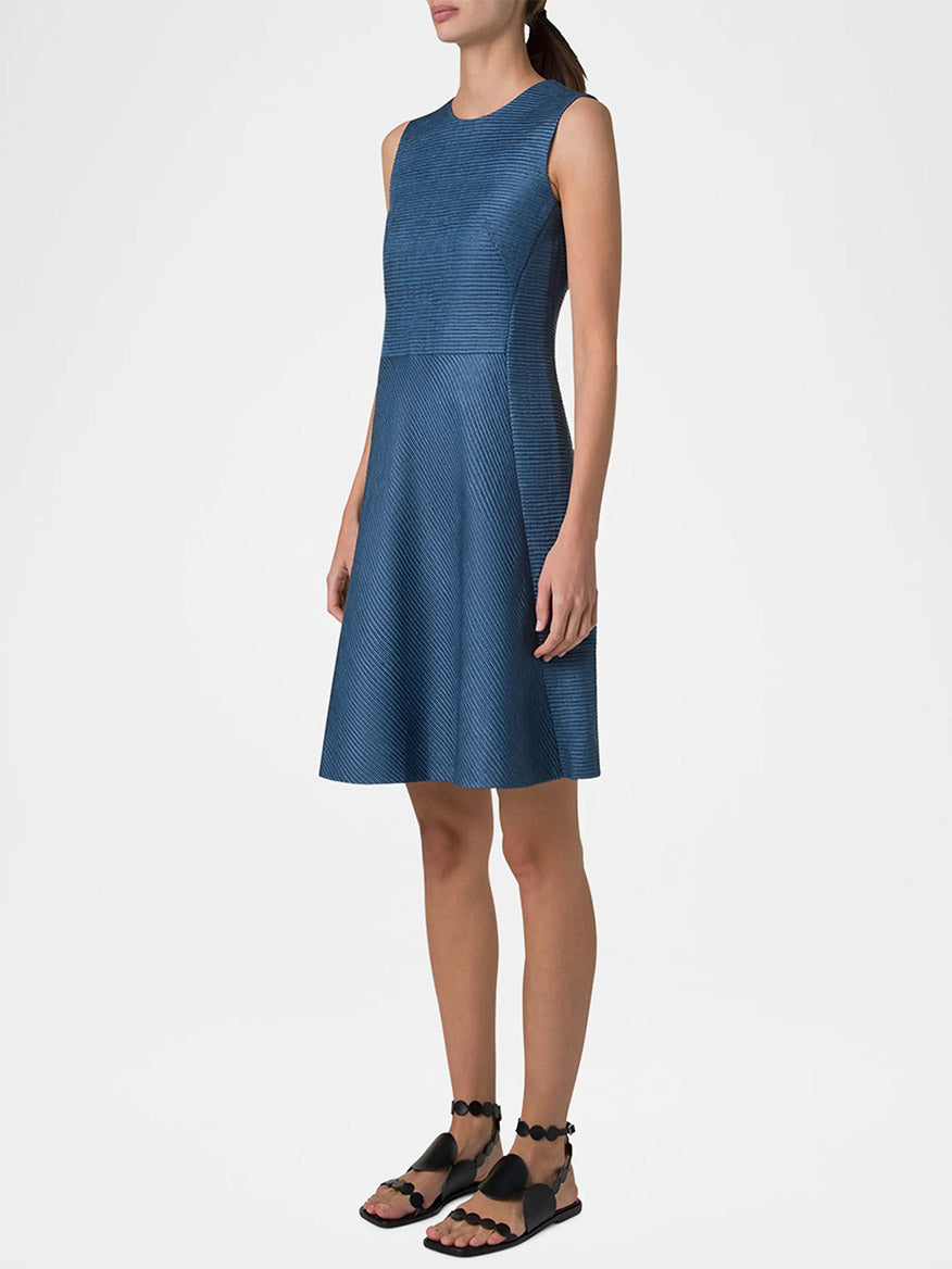 Dressed in the Akris Punto Sleeveless Silk Ottoman Shift Dress in Medium Blue and black sandals, a person stands gracefully against a plain backdrop.