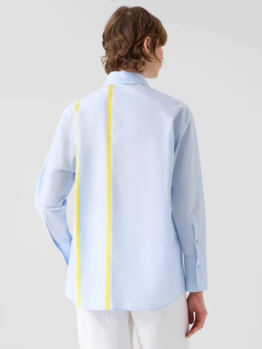 A person wears the Akris Punto Striped Cotton Chambré Shirt Blouse in Sky/Lemon/Cream, featuring a light blue relaxed fit and two vertical yellow back stripes, standing against a plain light background.