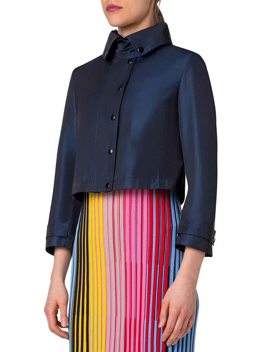 A person is wearing an Akris Punto Techno Water Resistant Satin Short Jacket in Ink over a pleated dress with vertical stripes in various colors including yellow, pink, red, and blue.