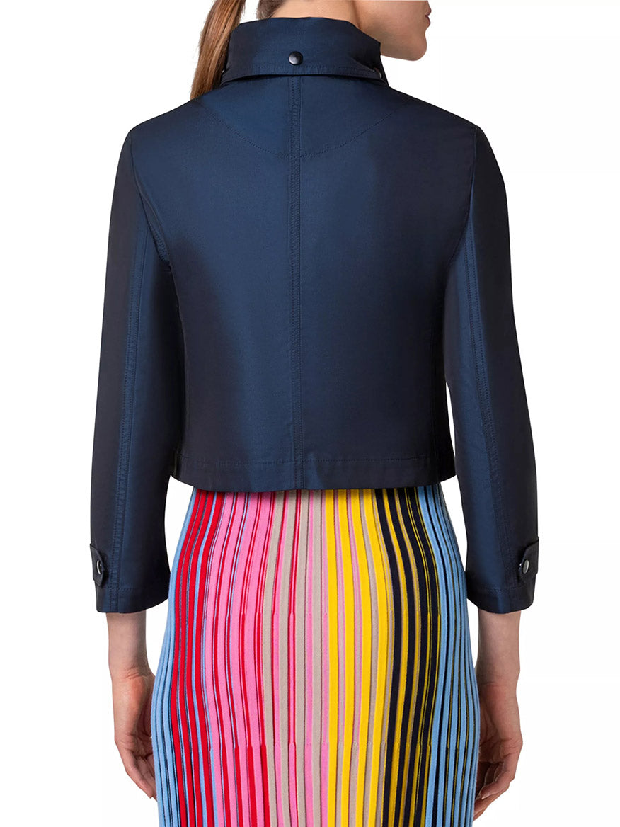 Rear view of a person wearing an Akris Punto Techno Water Resistant Satin Short Jacket in Ink with a snap-button collar and three-quarter sleeves, paired with a colorful, vertically striped skirt in red, blue, yellow, pink, and black shades.