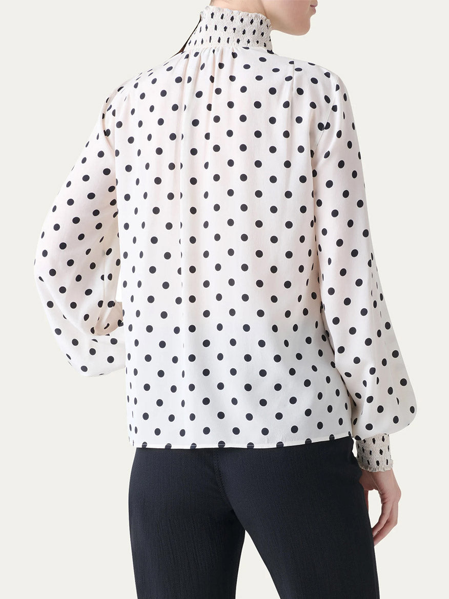 A person shown from the back wearing the Akris Punto Smocked Polka Dot Silk Blouse in Cream/Black with black pants.