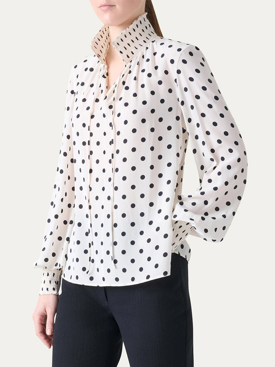 A person wearing the Akris Punto Smocked Polka Dot Silk Blouse in Cream/Black with black trousers, standing against a plain background.