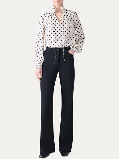 A person wearing the Akris Punto Smocked Polka Dot Silk Blouse in Cream/Black with high-waisted black flared pants, standing against a plain light background.