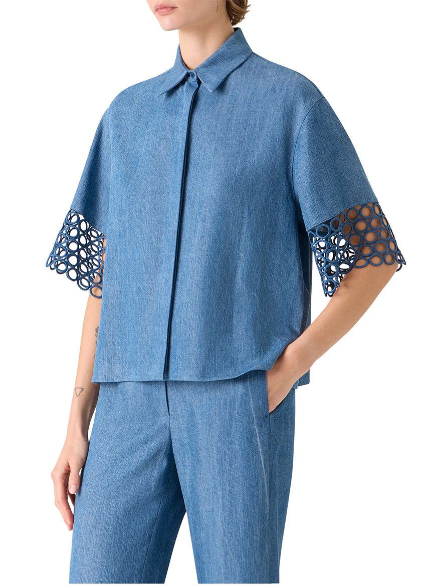 Wearing the Akris Punto Washed Denim Circle Lace Shirt in Medium Blue with open-circle lace on the elbow-length sleeves and matching denim pants, the person stands with one hand in a pocket.