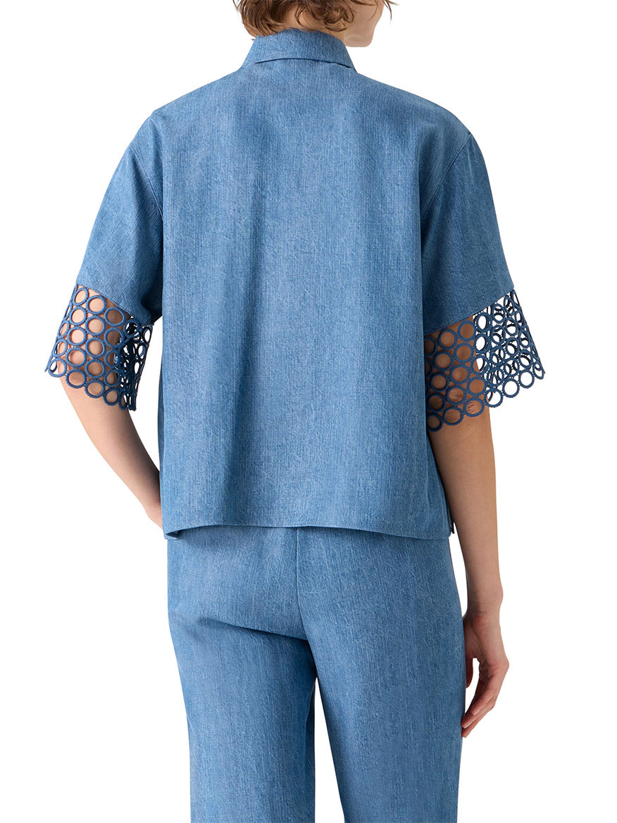 A person is shown from the back wearing the Akris Punto Washed Denim Circle Lace Shirt in medium blue, featuring open-circle lace cut-outs on the elbow-length sleeves, paired with matching blue pants.