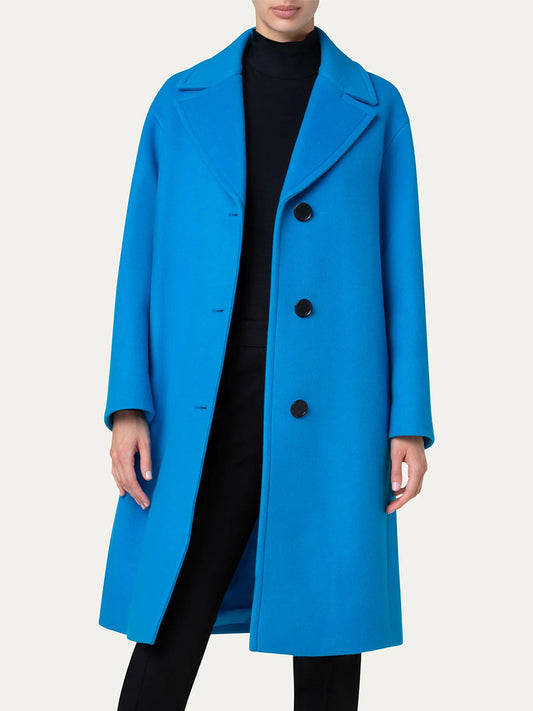 A person wearing an Akris Punto Single Breasted Wool Coat in Turquoise with black buttons over a black outfit, standing against a plain background.