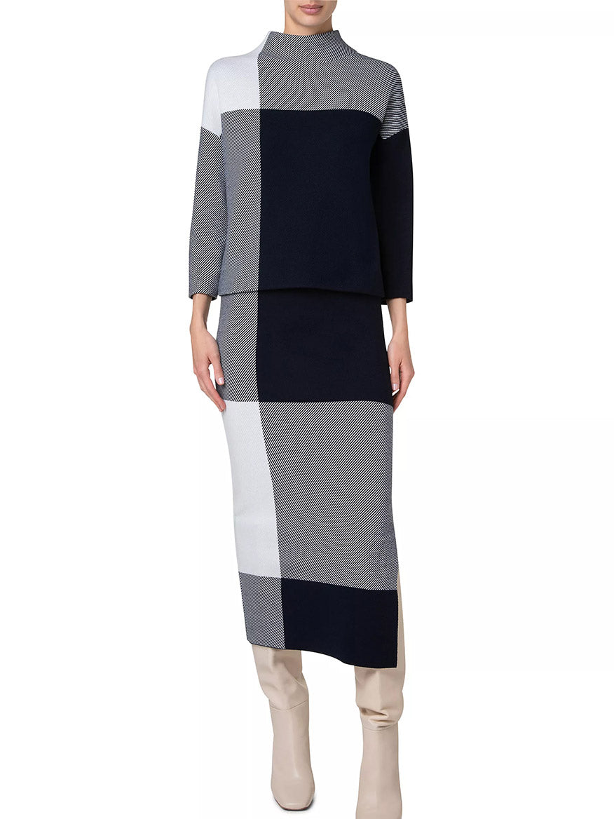 A person wearing a long-sleeve, color-blocked dress with black, white, navy, and gray squares paired with light-colored knee-high boots and the Akris Punto XL Gingham Merino Wool Mock Neck in Cream/Navy.