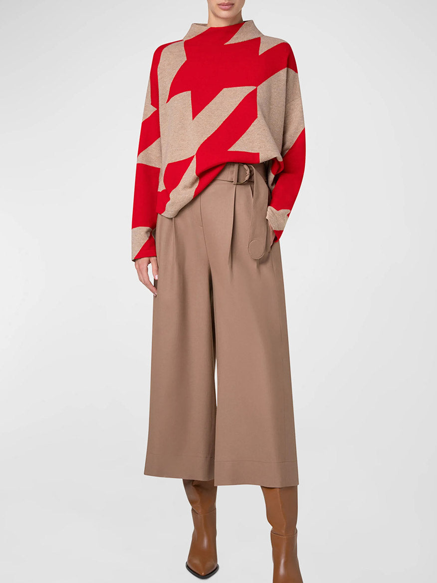 A person wearing an Akris Punto XL Houndstooth Wool/Cashmere Mockneck in Lipstick/Caramel, paired with beige wide-leg trousers and brown heeled boots, stands against a plain background.