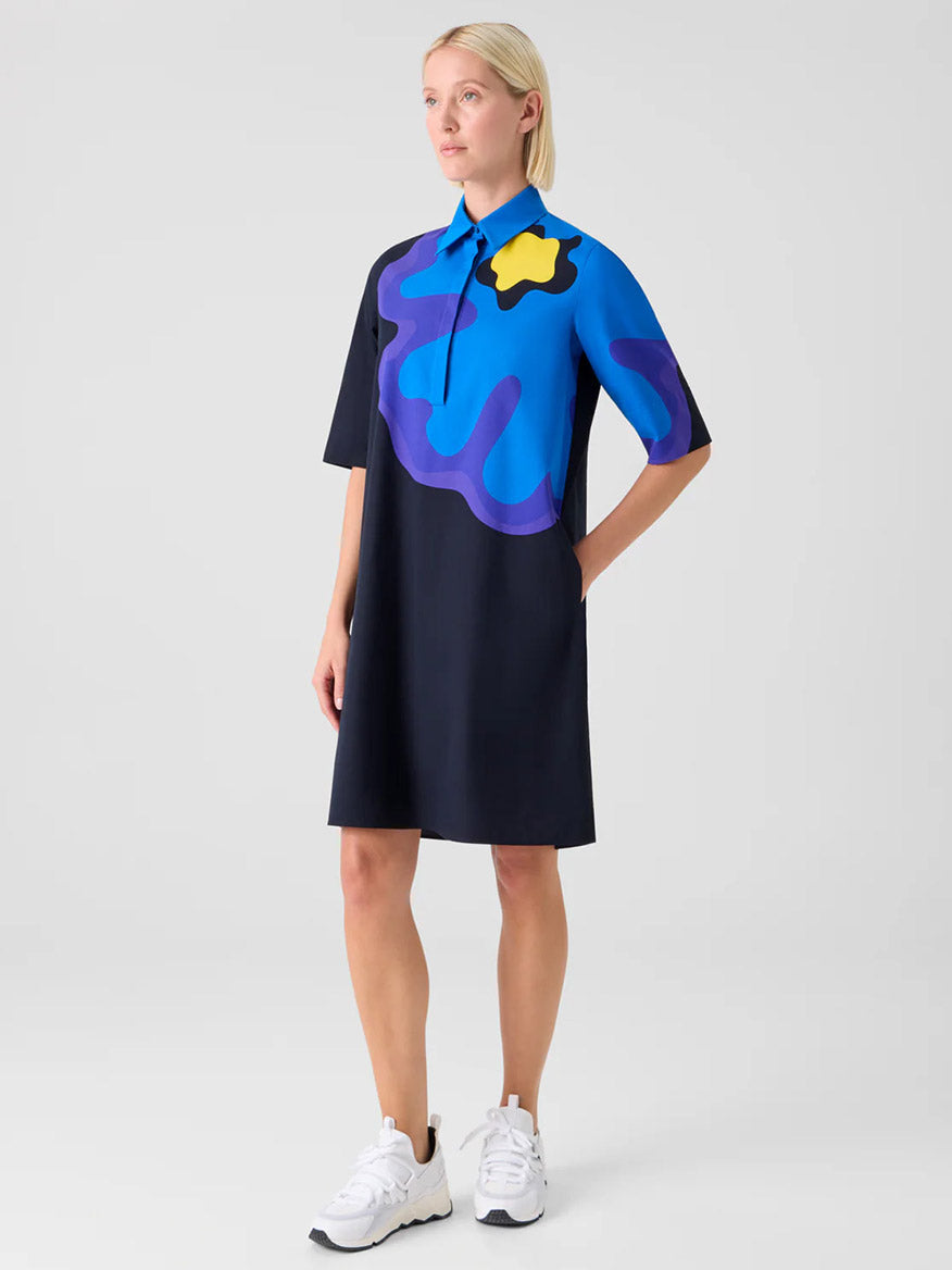 A woman stands against a plain background wearing the Akris Punto XL Pop Dot Flower Cotton Dress in Black Multi, paired with white sneakers and her hands in her pockets.