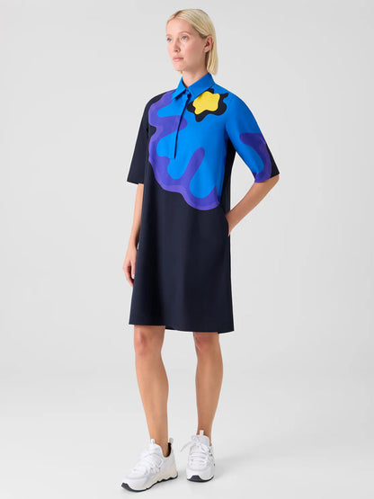 A woman stands against a plain background wearing the Akris Punto XL Pop Dot Flower Cotton Dress in Black Multi, paired with white sneakers and her hands in her pockets.