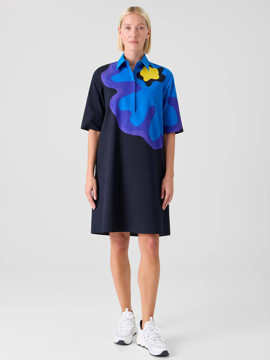 A woman faces forward wearing the Akris Punto XL Pop Dot Flower Cotton Dress in Black Multi, featuring bold blue, purple, and yellow patterns, styled with white sneakers against a plain background.
