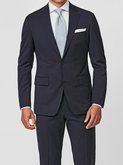 A man stands against a plain light background wearing the Atelier Munro Midnight Blue Twill 4-Season Wool Stretch Suit, paired with a light blue tie, white dress shirt, and white pocket square.