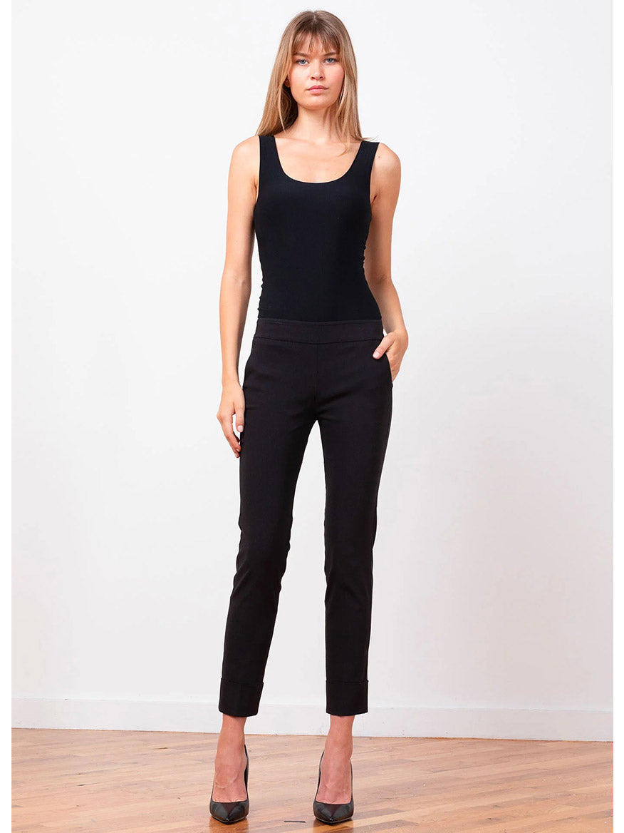 A woman stands against a white background wearing a black sleeveless top, the Avenue Montaigne Ben Signature Straight Leg Pant in Black made of stretch fabric, and black high heels.