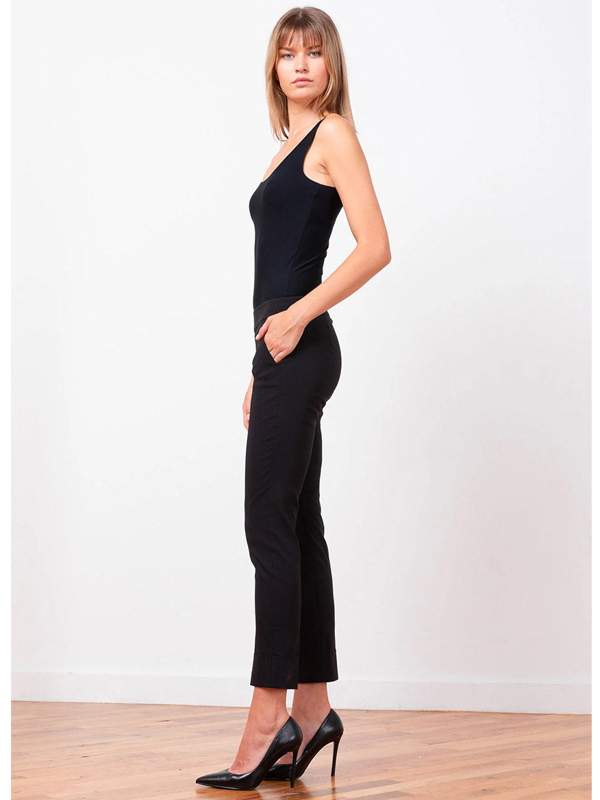 A woman stands against a plain white background, dressed in the sleek Avenue Montaigne Ben Signature Straight Leg Pants in black and a matching sleeveless top. She has one hand in her pocket and confidently looks at the camera.