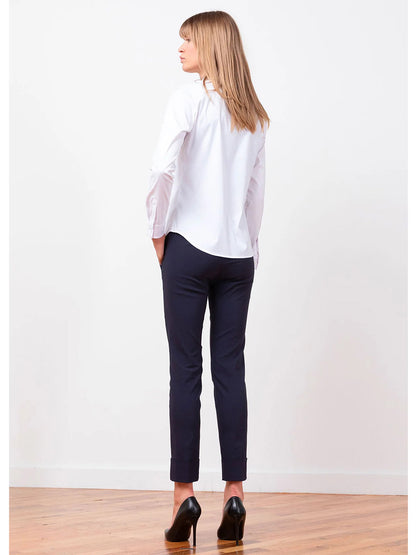 A woman with blonde hair stands facing away on a hardwood floor, wearing a white button-up shirt, Avenue Montaigne Ben Signature Straight Leg Pant in Navy, and black high heels. The plain white background accentuates her elegant look, reminiscent of the chic style found on Avenue Montaigne.