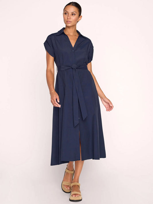 A woman in a navy blue Brochu Walker Fia Belted Dress in Navy with a tie waist, standing against a light background, wearing beige wedge sandals.
