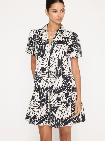 A woman is wearing a short-sleeve, button-up Brochu Walker Havana Mini Dress in Palm Combo. She has her hair pulled back and is standing against a plain background.