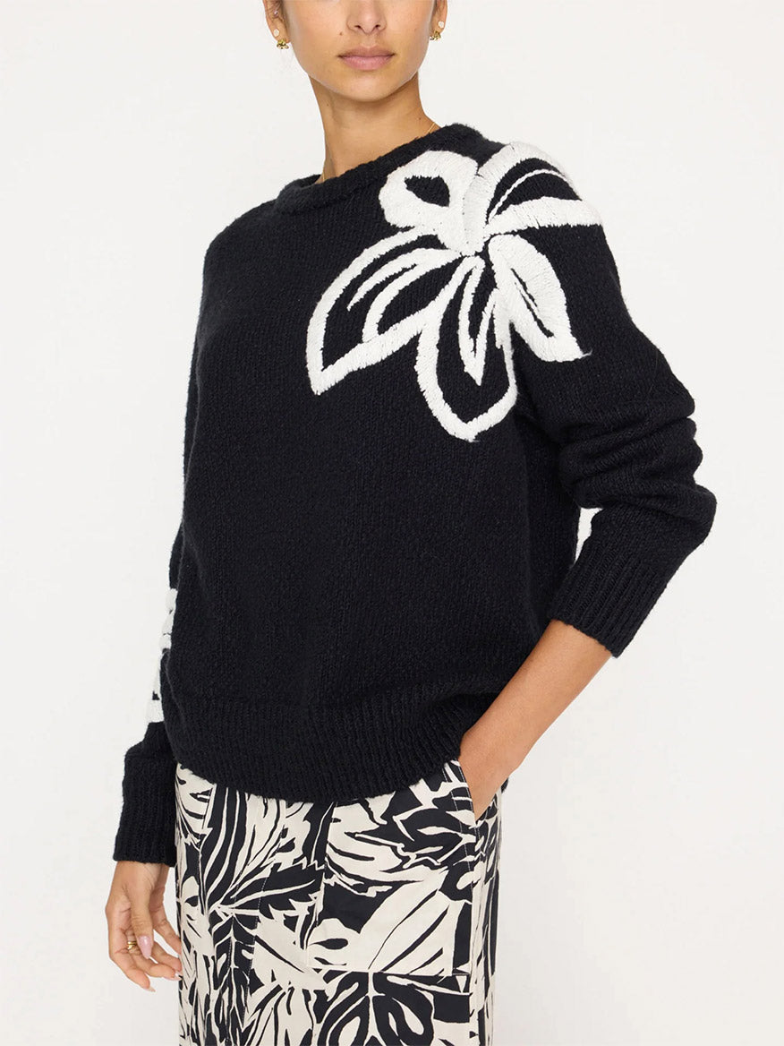 Person wearing a black Brochu Walker Hibiscus Embroidered Sweater in Black Onyx with White featuring a white floral design on the shoulder, paired with a black and white floral skirt.