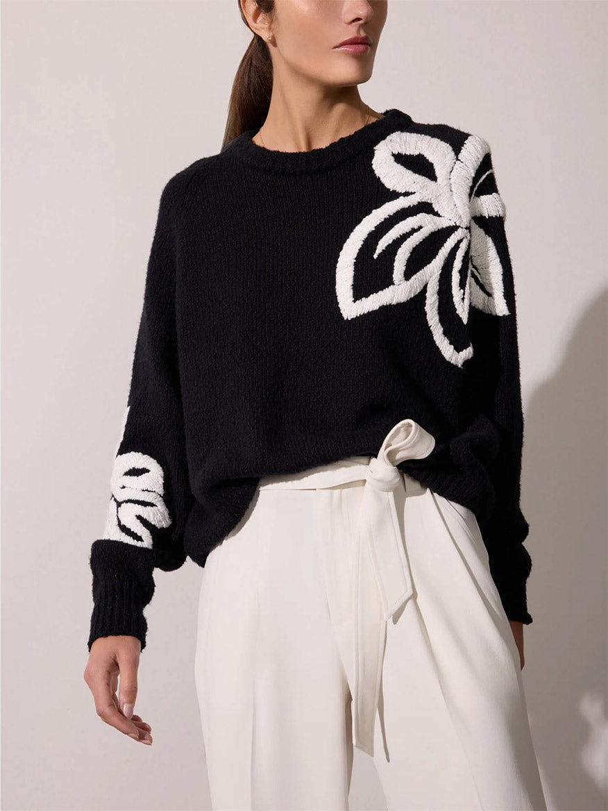 A person is wearing a black Brochu Walker Hibiscus Embroidered Sweater in Black Onyx with White with large white floral designs on the shoulder and wrist, paired with high-waisted white trousers tied with a white belt.