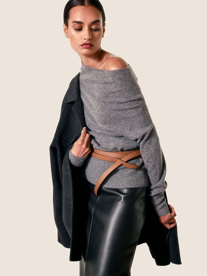 A person is elegantly posing against a plain background in a chic Brochu Walker Lori Off Shoulder Sweater in Flint Melange, with a dark coat draped over one shoulder and a black leather skirt accented by a tan belt.